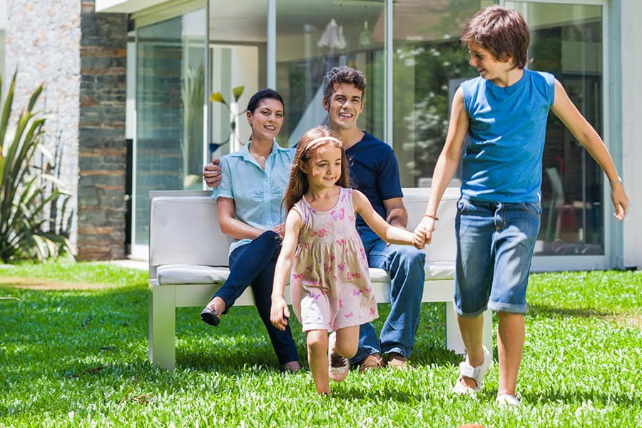 Homeowner’s Insurance in Vermont - Brother Holding Sisters Hand as They Play in the Yard of Their House with Their Parents Smiling and Watching them from a Bench in the Yard